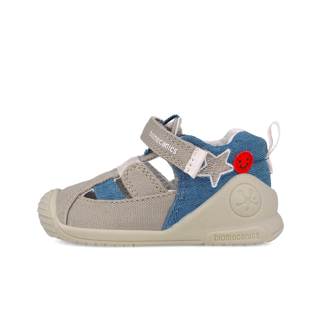 Children's sneaker with beige and blue denim fabric, a Velcro strap, star patches, and a red smiley patch on the heel; beige sole labeled Biomecanics.